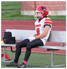 A person wearing a red football helmet and white jersey with the number 5 is sitting on a bench next to a black bag on a sports field. Description generated by AI