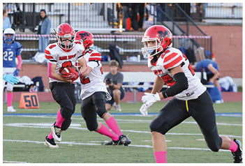 Players in red and white uniforms are running and preparing to tackle players in blue and white uniforms during a football game on a field with spectators in the background. Description generated by AI