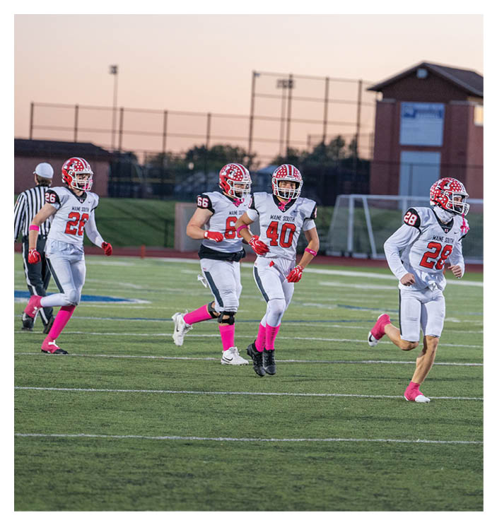 Four people in white football uniforms with red helmets and pink socks are running on a green field during a game at dusk. Description generated by AI