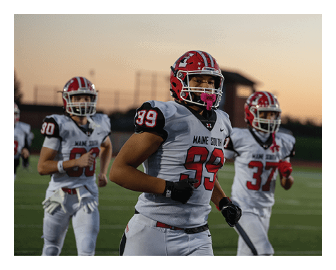 Three people in football uniforms with red helmets and white jerseys are running on a field during sunset. Description generated by AI