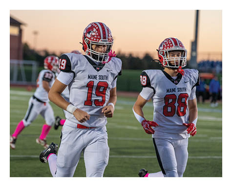 People in football uniforms with helmets and pink accessories are running on a field during sunset. Description generated by AI