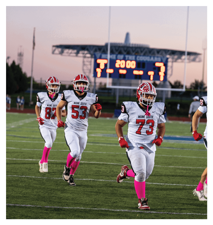 Four people in football uniforms with pink socks are running on a field with a scoreboard in the background showing the start of a game. Description generated by AI