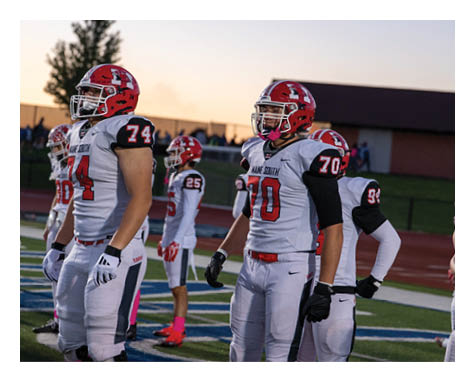 Several people wearing football uniforms and helmets stand on a field during sunset. Description generated by AI