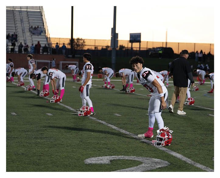 People in football uniforms are stretching on a field at sunset with helmets placed on the ground. Description generated by AI