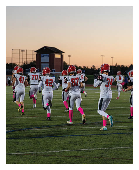 A group of people wearing white sports uniforms with red helmets and pink socks are running on a green football field during sunset. Description generated by AI