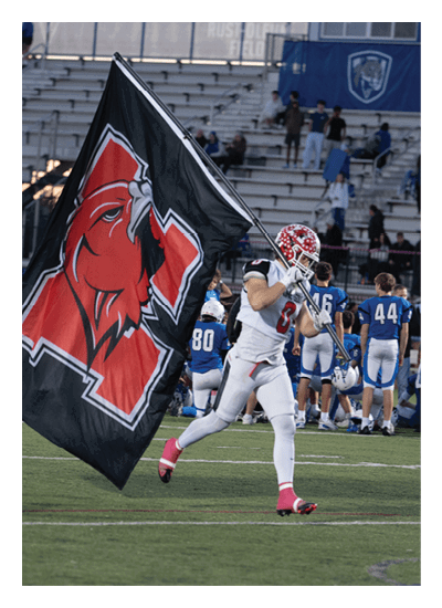 A person in a white football uniform with pink cleats is running on a football field while holding a large black flag with a red hawk logo and a red letter N. Description generated by AI
