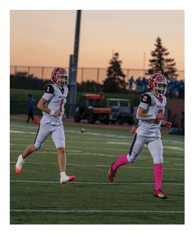 Two people in football uniforms and helmets are running on a field during sunset with a few people and equipment visible in the background. Description generated by AI