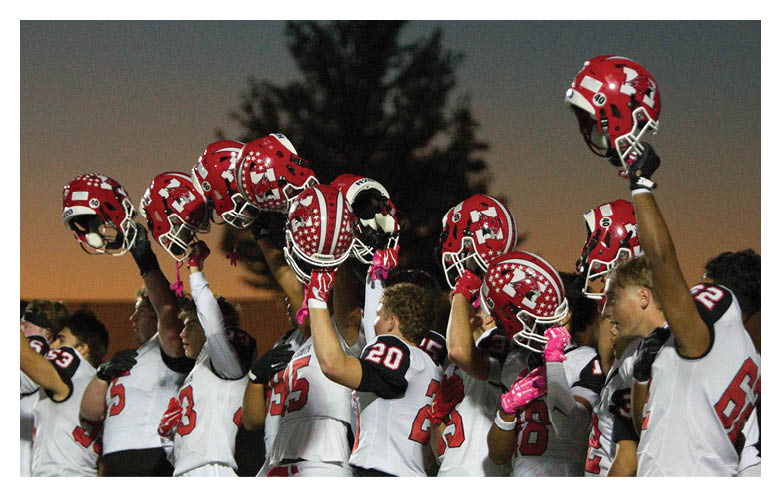 A group of people wearing sports uniforms raise red helmets with white patterns in the air during an outdoor event at dusk. Description generated by AI
