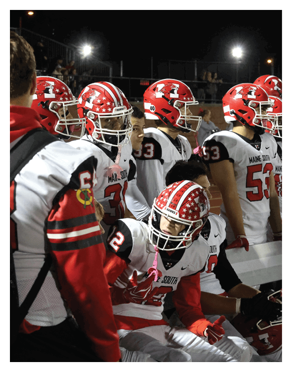 Several people wearing red and white football uniforms and helmets are gathered closely together, with one person sitting and gesturing while others stand nearby. Description generated by AI