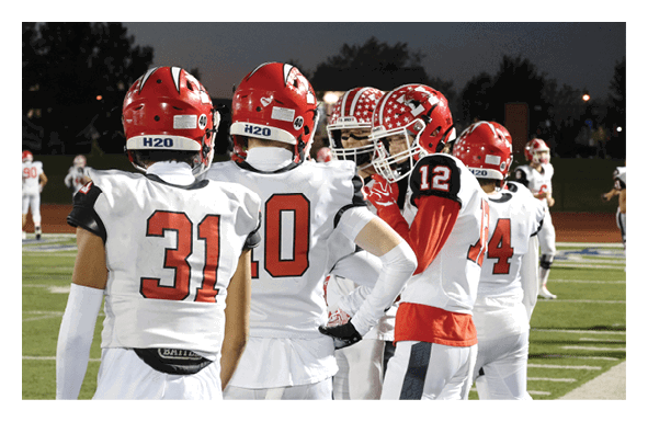 Four people wearing football helmets and uniforms stand on a field during a game at dusk. Description generated by AI