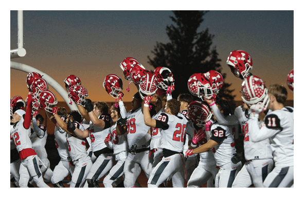 A group of people wearing white sports uniforms with red helmets raised in the air stand together on a football field at sunset. Description generated by AI