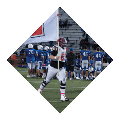 People in blue football uniforms are gathered on a field near the sidelines while a person in a white and red uniform stands in the foreground holding a flag. Description generated by AI