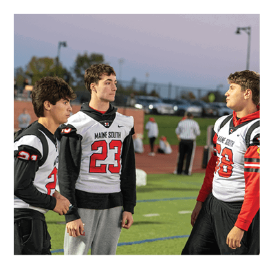 Two people wearing football jerseys and hoodies are standing on a sports field having a conversation with a referee and other people in the background. Description generated by AI