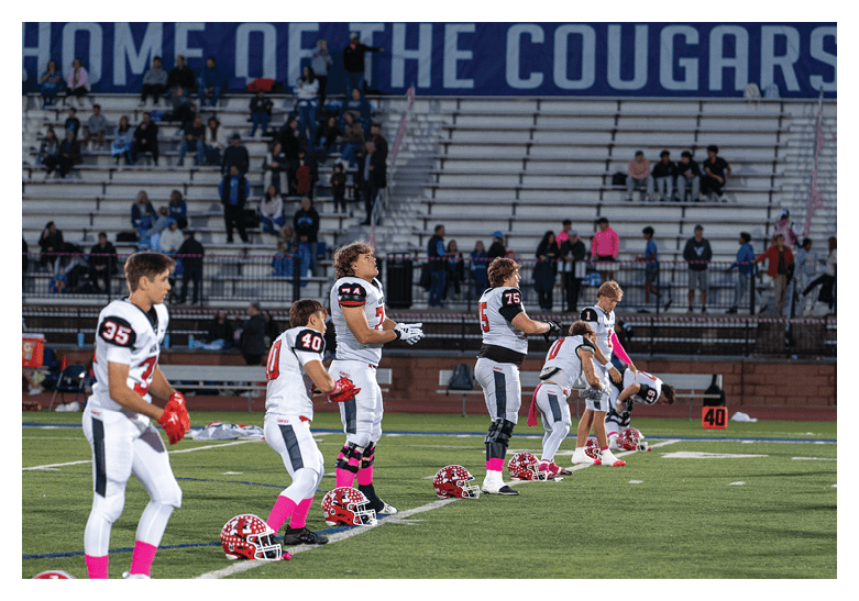 Several people in football uniforms with pink socks are kneeling on a field next to their helmets during a moment of pause, with empty bleachers and a banner reading “HOME OF THE COUGARS" in the background. Description generated by AI