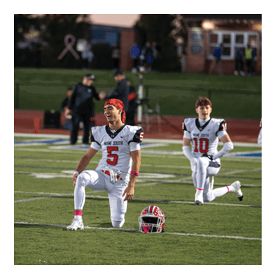 Two people in football uniforms kneel on a field with helmets placed in front of them during what appears to be a pre-game or halftime moment. Description generated by AI