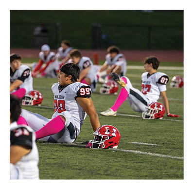 People in football uniforms with pink socks are sitting on a field stretching with their helmets placed on the ground nearby. Description generated by AI