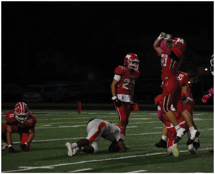 People in football uniforms are on a field at night with one person in red jumping to catch a ball while others watch or kneel nearby. Description generated by AI