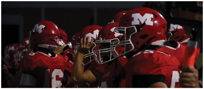 A group of people wearing red football helmets and uniforms with a white “M" logo are gathered closely together, with one person raising a hand. Description generated by AI
