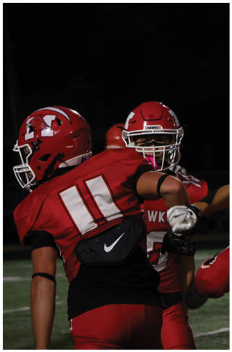 Two people wearing red football uniforms and helmets are on a field at night, with one person facing away and the other looking toward them. Description generated by AI