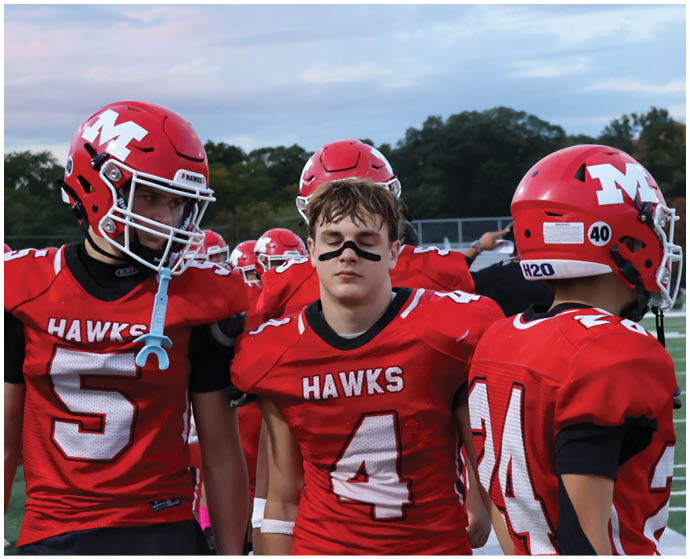 Three people wearing red football uniforms with “HAWKS" on the front stand together on a field with helmets, one person has black face paint under their eyes. Description generated by AI