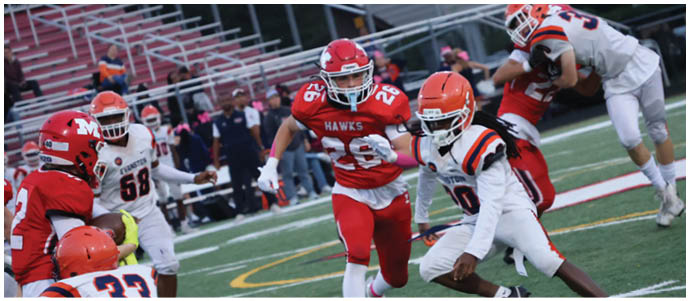 People in football uniforms are actively playing on a field with one person running while others are preparing to tackle or block. Description generated by AI