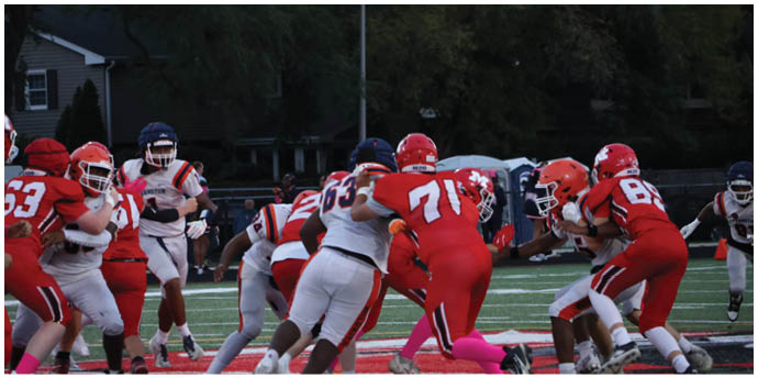 People wearing football helmets and uniforms are engaged in a play on a grassy field with trees and houses in the background. Description generated by AI
