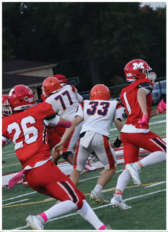 Several people wearing football uniforms in red and white are actively playing on a green field with trees and vehicles in the background. Description generated by AI