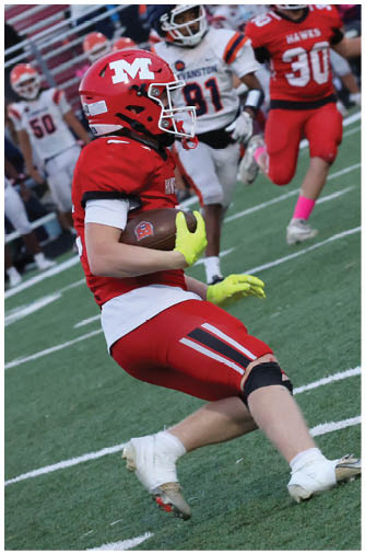 A person in red football gear with a helmet and neon yellow gloves is running with a football on a field while other players in red and white uniforms chase behind. Description generated by AI