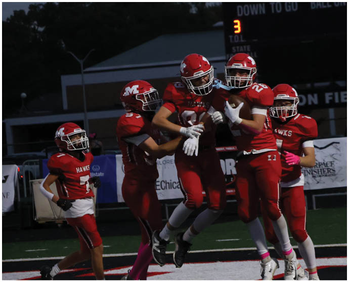 Five people in red football uniforms celebrate on a field with a scoreboard showing the score and time in the background. Description generated by AI
