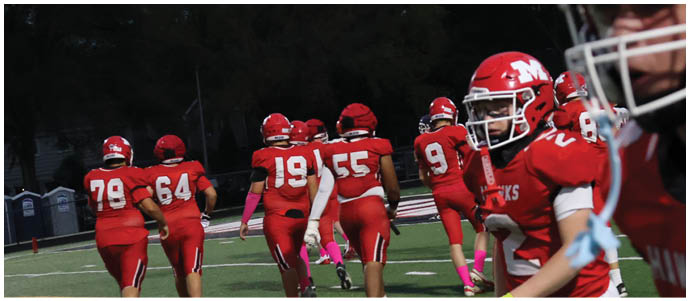 People wearing red football uniforms and helmets are gathered on a field with trees and portable toilets visible in the background. Description generated by AI