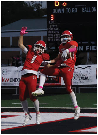 Two people in red football uniforms are celebrating on a football field with a scoreboard showing the score and time in the background. Description generated by AI