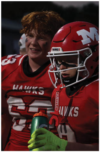 Two people wearing red football uniforms with “HAWKS" on the jerseys are standing together, one smiling and the other holding a green water bottle while wearing bright green gloves and a helmet. Description generated by AI