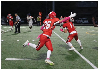 Players in red and white football uniforms are actively engaged in a nighttime game on a field with a referee observing the play. Description generated by AI