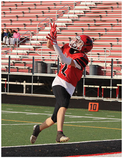 A person in a red football uniform and helmet is jumping to catch a football near the end zone on a field with empty bleachers in the background. Description generated by AI