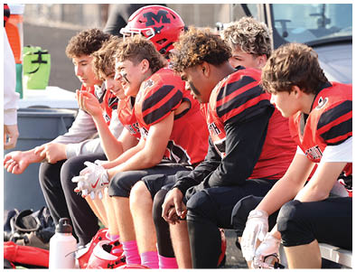 People wearing red and black football uniforms are sitting closely together on a bench, some with gloves on and one person holding a football. Description generated by AI