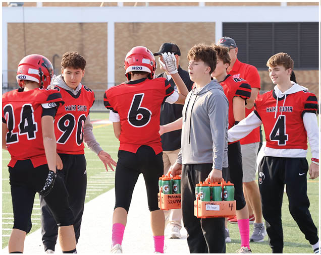 People wearing red football uniforms and helmets are gathered on a field while others hold water bottles and interact. Description generated by AI