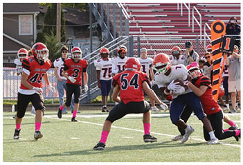 Children playing a football game on a field with one player being tackled while others approach, and a group of players and coaches stand on the sidelines near empty bleachers. Description generated by AI