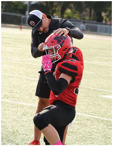 An adult wearing a black jacket and cap adjusts the helmet of a person in a red football uniform on a sports field. Description generated by AI