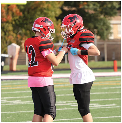 Two people wearing red football jerseys and helmets stand facing each other on a football field, with one adjusting the other's gloves. Description generated by AI