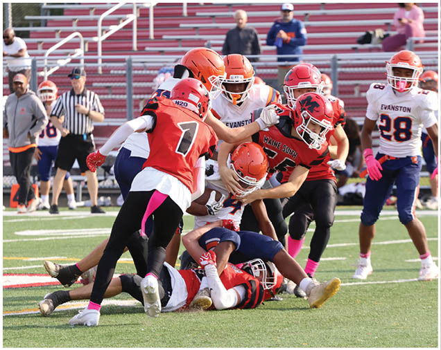 Several people wearing football helmets and uniforms are engaged in a tackle on a football field with empty bleachers in the background. Description generated by AI