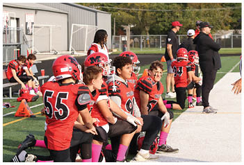 A group of people wearing red and black football uniforms sit on a bench on the sidelines while a person in a black and white striped referee uniform stands nearby on a sports field. Description generated by AI