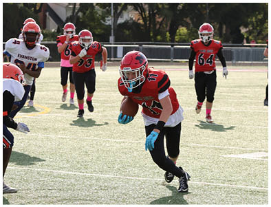 A group of people wearing red and black football uniforms are playing on a sunlit field with one person running with the ball while others prepare to defend or support. Description generated by AI