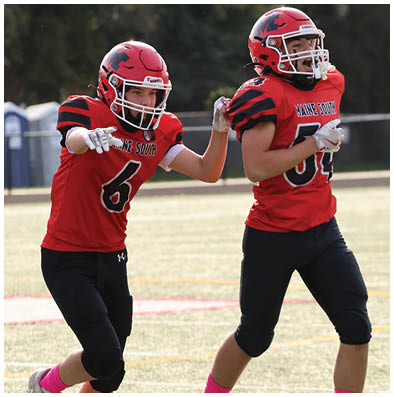 Two people in red football uniforms and helmets are on a field with one pointing forward and the other running beside them. Description generated by AI