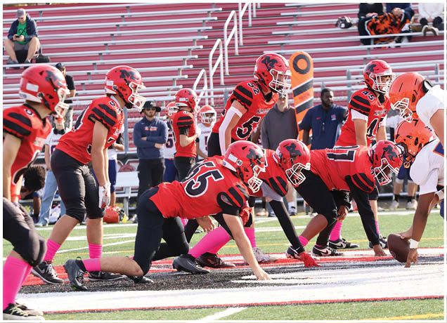 A group of people in red and black football uniforms are lined up on a field facing a group in white and orange uniforms, preparing for a play. Description generated by AI
