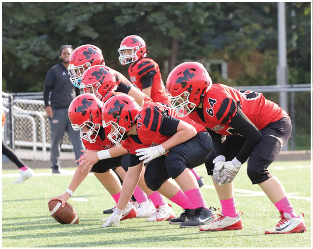 Five people in red and black football uniforms with helmets are lined up on a grassy field preparing for a play, with one person holding a football ready to snap it. Description generated by AI