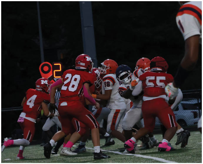A group of people wearing football uniforms are engaged in a play on a field under dim lighting with a down marker showing the number two in the background. Description generated by AI