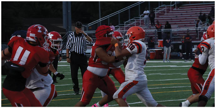 People in football uniforms are engaged in a play on a field with a referee observing nearby and empty bleachers in the background. Description generated by AI