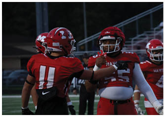 People wearing red football uniforms are interacting on a field with bleachers and a dark background. Description generated by AI