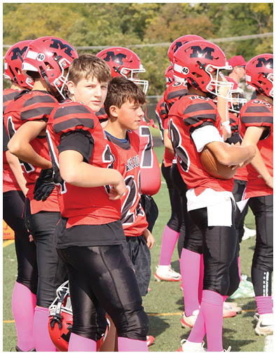 A group of people wearing red and black football uniforms with pink socks stand on a grassy field, some holding helmets and one holding a football. Description generated by AI