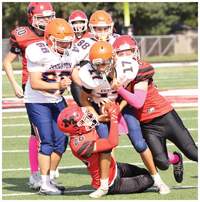 Several people wearing football helmets and uniforms are engaged in a tackle on a grassy field during a game. Description generated by AI